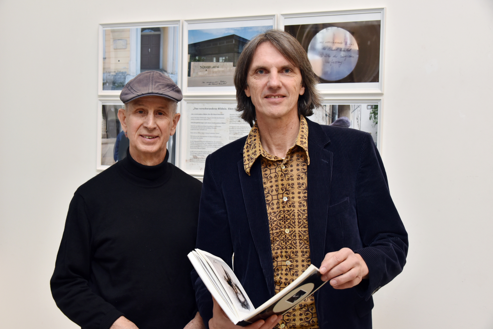 Horst Hoheisel und Andreas Knitz stehen vor einer Wand an der gerahmte Bilder angebracht sind. Der rechts stehende Andreas Knitz hält ein Buch in er Hand. Beide schauen in die Kamera.