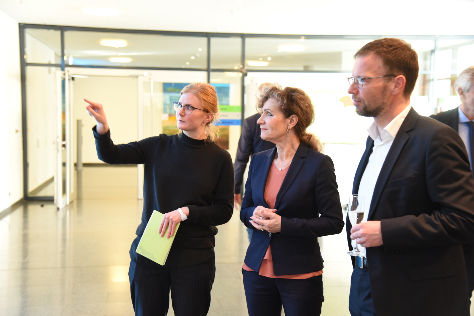 Links zeigt eine schwarz gekleidete Person der rechts von ihr stehenden Frau und dem Mann ein Ausstellungsstück, das nicht im Bild zu sehen ist.