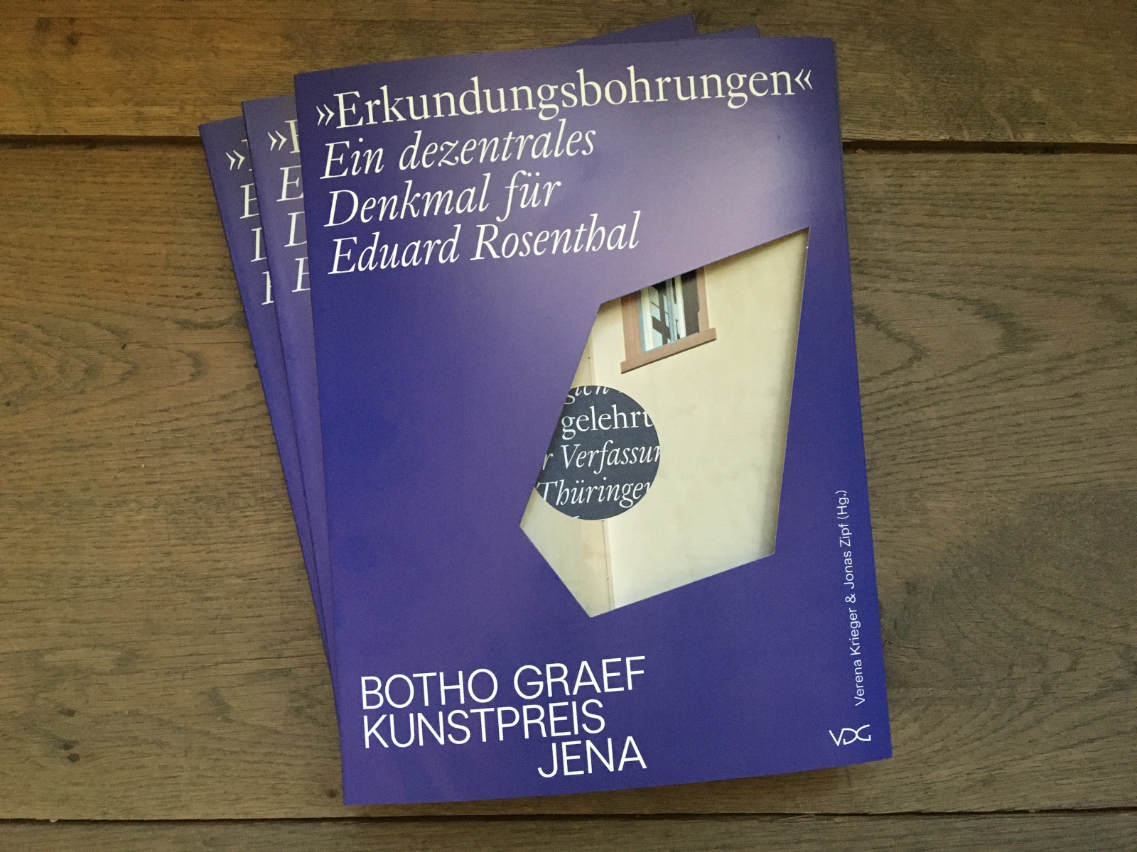 Drei Exemplare des Katalogs, ein Buch in dunkelblauem Umschlag mit Stanzungen durch die Buchdecke, liegen gestapelt auf einem Holzboden.