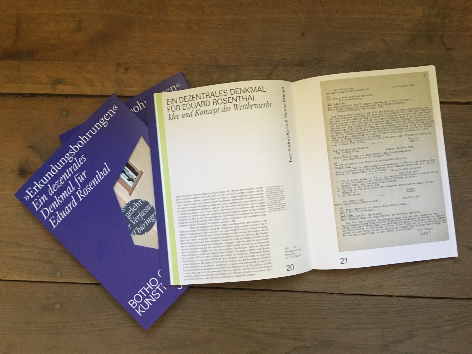 Drei Exemplare des Katalogs, ein Buch in dunkelblauem Umschlag mit Stanzungen durch die Buchdecke, liegen aufgeschlagen auf einem Holzboden.