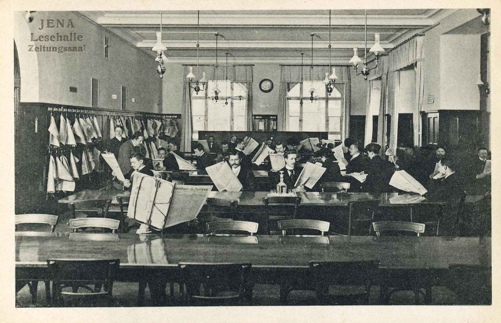 Blick in den Zeitungssaal der Lesehalle. An hintereinander aufgereihten Tischen sitzen mehrere Personen mit Zeitungen in der Hand.