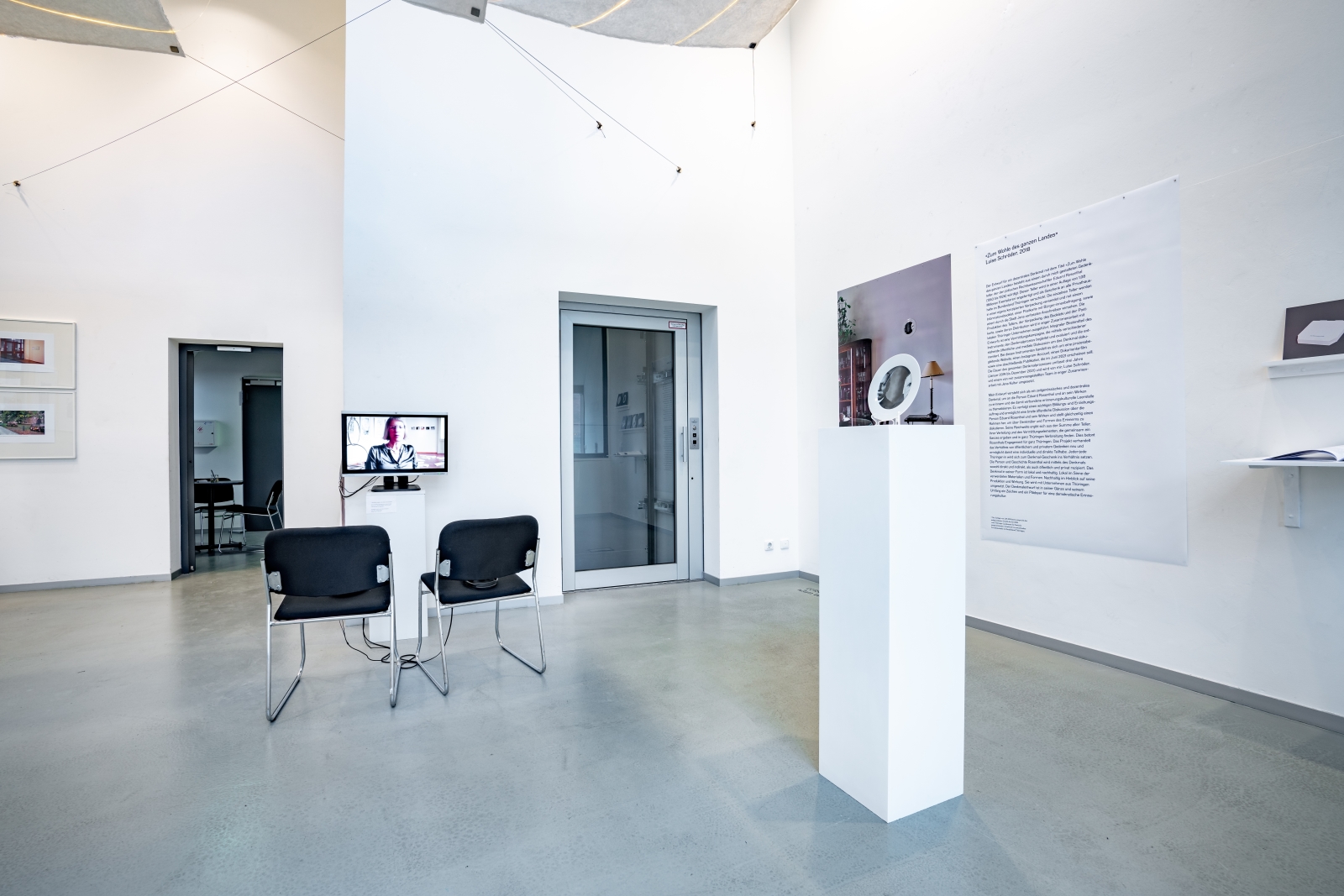Der obere Ausstellungsraum des Jenaer Kunstverein: In dem hellen Raum ist links der Entwurf zum Denkmal-Teller von Luise Schröder, in der Bildmitte ein Bildschirm mit einem Videointerview und zwei Stühlen zu sehen.